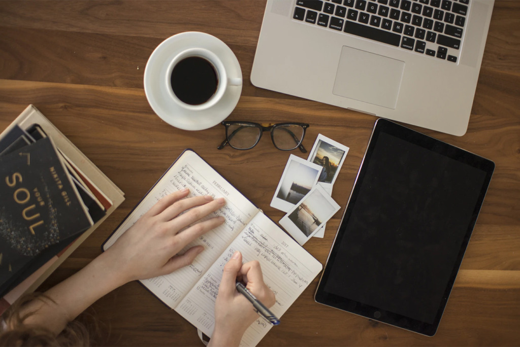 Persoon die in een notitieboekje schrijft aan een houten bureau met een laptop, tablet, koffie, bril, boeken en foto's, allemaal hulpmiddelen om hun SEO-blogstrategie te perfectioneren.