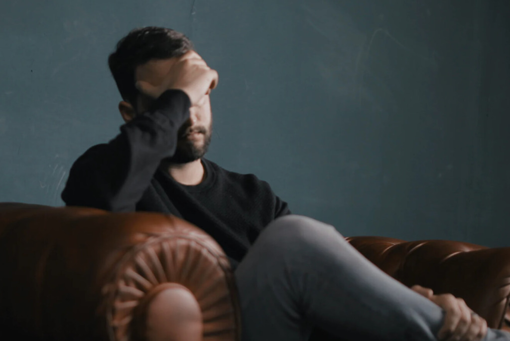 Een persoon die op een bruinleren bank zit, zijn hoofd in de hand laat rusten, contemplatief kijkt en waarschijnlijk worstelt met deadlinestress.