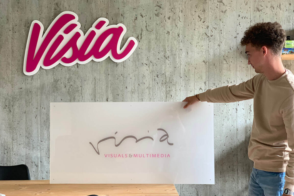 Een persoon die een bord vasthoudt met de tekst "Visia Visuals & Multimedia" staat naast een muur versierd met het rebrandinglogo van "Visia".