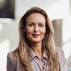 Een vrouw met lang blond haar in een beige blazer kijkt naar de camera en straalt het vertrouwen uit van Straight-Line Leadership, met een onscherpe achtergrond van een lichte binnenruimte.