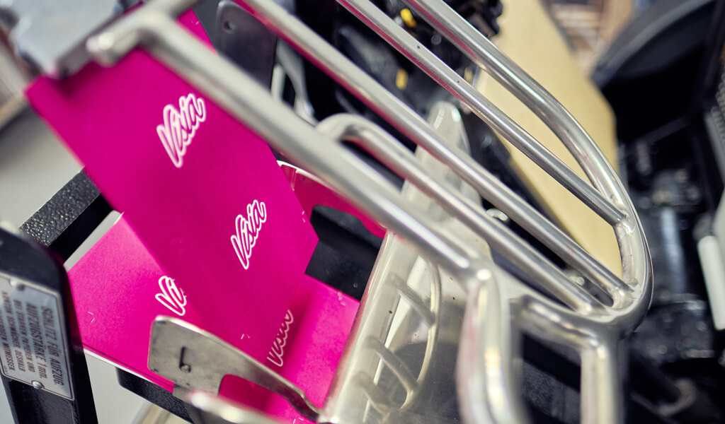 Een close-up van een drukpers met roze papier met witte "Veloz"-tekst die door metalen rollen wordt gevoerd, toont indrukwekkend drukwerk.