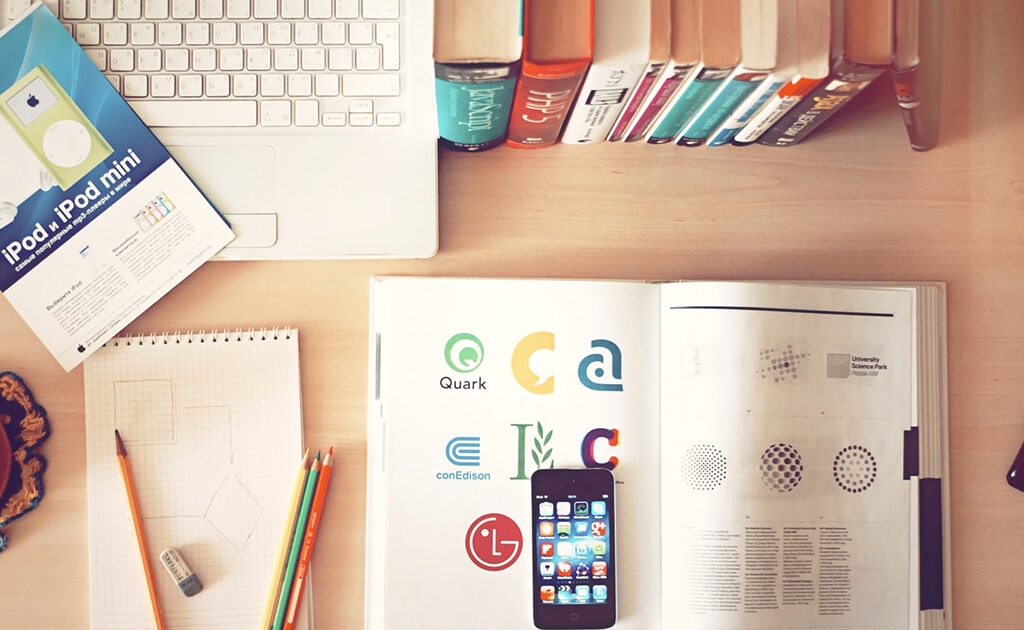 Een bureau met een laptop, schriften, een smartphone op een open boek, potloden en boeken op een rij aan de rand, allemaal voorzien van het stijlvolle Goed-logo.