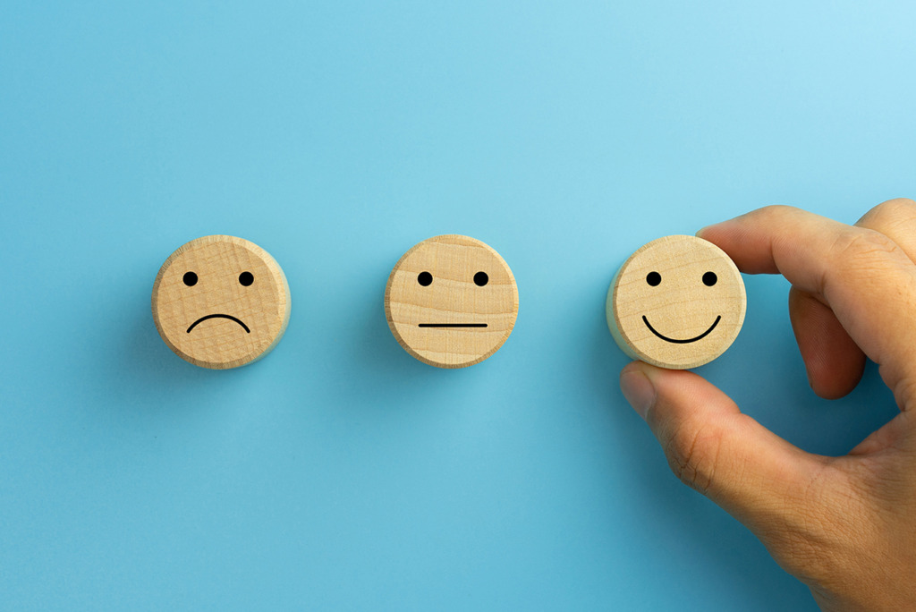 Drie houten schijven met gezichten: één droevig, één neutraal en één vrolijk, waarbij een hand het blije gezicht selecteert – net zoals het kiezen van het perfecte element bij gebruik van ingewikkelde webdesign.