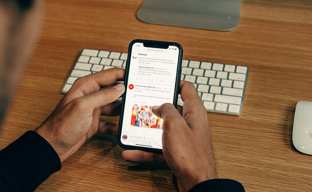 Persoon die op een smartphone aan een houten bureau met een wit toetsenbord en muis op Twitter surft en af en toe enkele nuttige Twitter-tips leest om zijn of haar sociale media-ervaring te verbeteren.