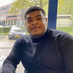 Een man in een zwarte coltrui zit aan een tafel in een buitenomgeving met bomen en een gebouw op de achtergrond, met een Computerkruis in zijn hand.