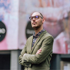Een man in een groene blazer en bril staat buiten met de armen over elkaar, naar boven kijkend, misschien met een beeld van de toekomst van Heerlen. Je kunt hem bijna 'Mijn Stad' horen fluisteren terwijl hij de omgeving in zich opneemt.