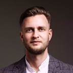 Een man met kort haar en een baard, gekleed in een wit overhemd en een blazer met patronen, staat zelfverzekerd tegen een effen donkere achtergrond, klaar om te gedijen.