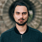 Buiten staat een man met lang haar en een baard, met een stenen boog op de achtergrond die de grens markeert.