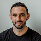 Een man met kort krullend haar en een baard, gekleed in een zwart Presuro-shirt, glimlacht naar de camera tegen een effen achtergrond.