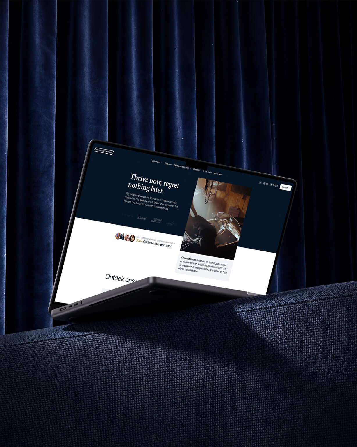 Op een donkerblauwe bank met een gordijn op de achtergrond staat een dunne laptop open; op het scherm is een website te zien — zijn doelgerichte uitstraling belichaamt de principes van Straight-Line Leadership.