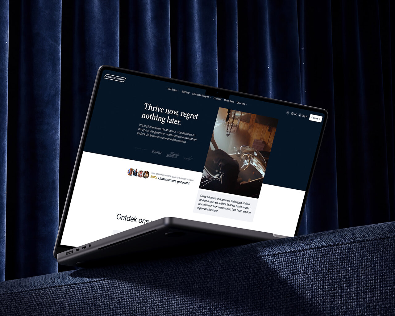Op een donkerblauwe bank met een gordijn op de achtergrond staat een dunne laptop open; op het scherm is een website te zien — zijn doelgerichte uitstraling belichaamt de principes van Straight-Line Leadership.