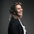 Een vrouw met blond haar, gekleed in een donkere blazer en witte blouse, glimlacht tegen een donkere achtergrond en straalt een sfeer van L'Authentique-elegantie uit.