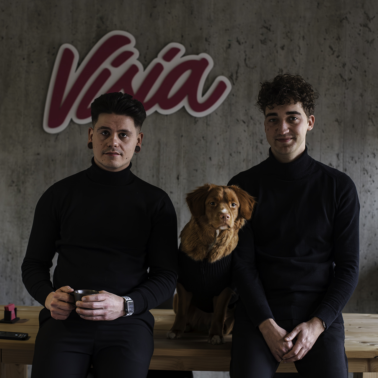 Twee mannen in zwarte truien en een bruine hond met een zwarte trui zitten op een bankje, poserend met een bordje 'Over ons' erachter.