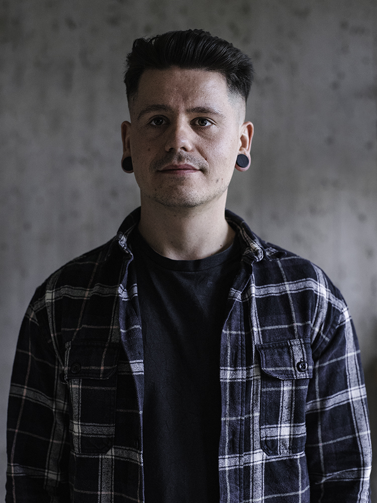 Een man met kort haar, gekleed in een zwart-wit geruit overhemd, staat met zijn gezicht naar de camera voor een grijze achtergrond.