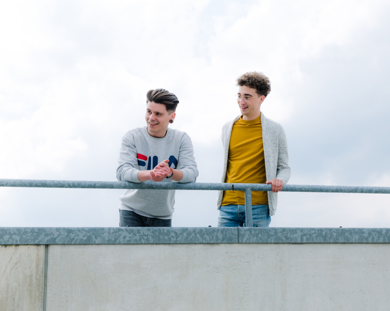 Twee jonge mannen, de een in een grijs sweatshirt en de ander in een geel overhemd, staan glimlachend op een dak met een blauwe lucht achter hen. Deze momentopname legt hun levendige energie vast en geeft een indicatie van wie ze zijn – over ons.