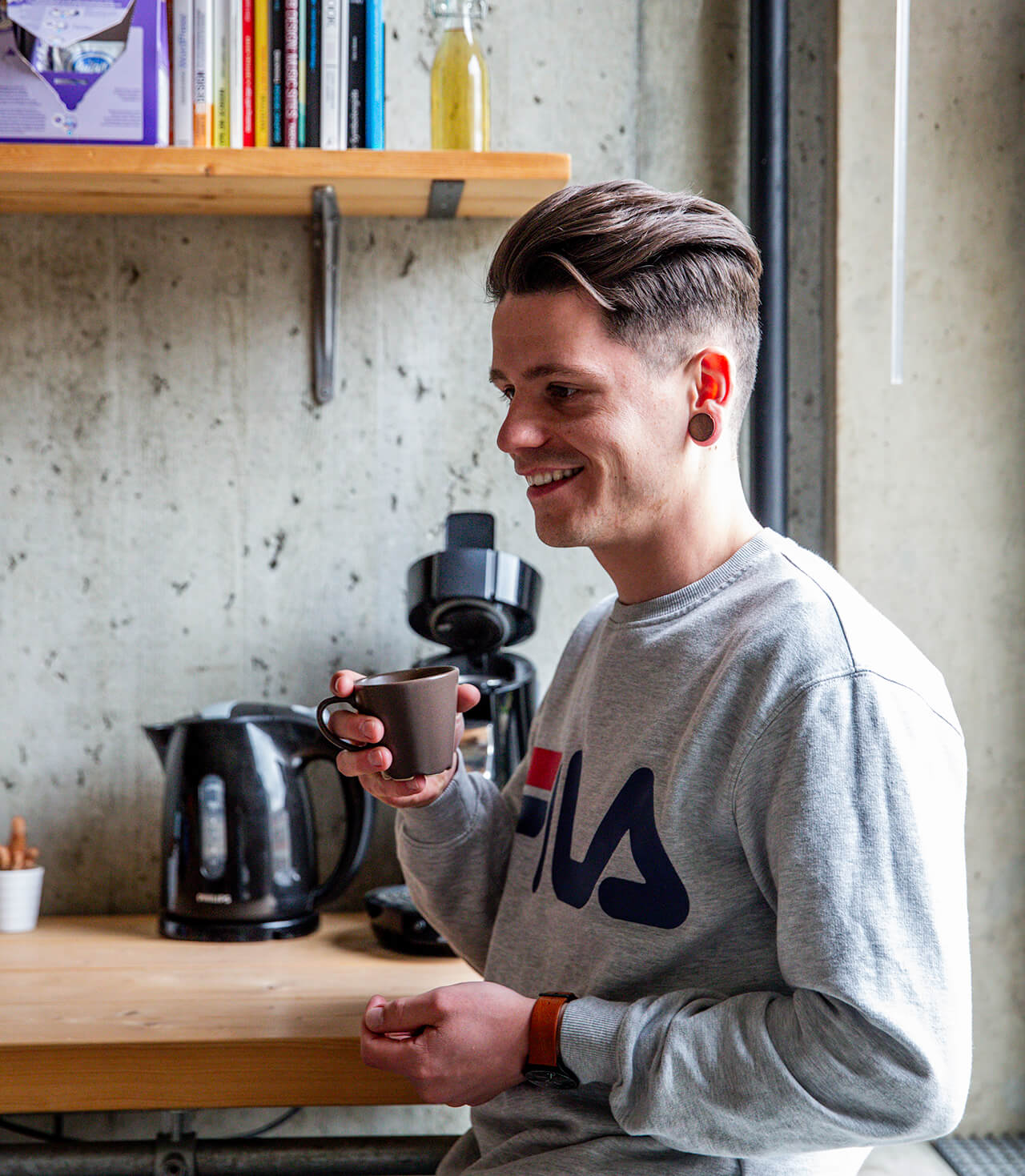 Man met kort haar, gekleed in een sweatshirt en koptelefoon, glimlachend terwijl hij een koffiemok in de keuken vasthoudt. Over ons sectie zou meer details bieden.