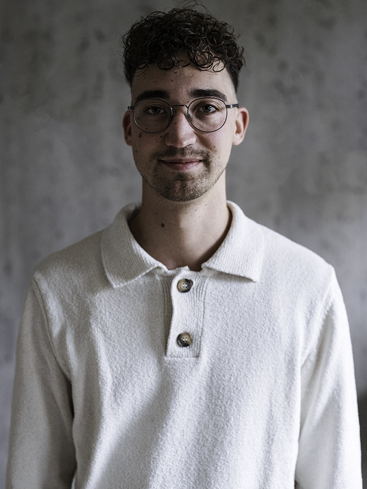 Een persoon met krullend haar en een bril, gekleed in een overhemd met witte kraag, staande tegen een effen achtergrond.