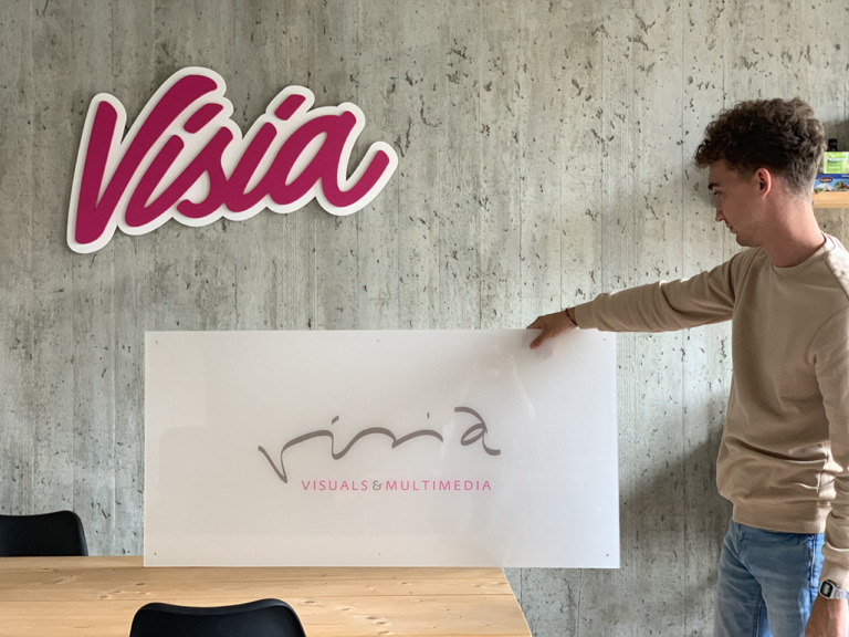 Een persoon houdt een Visia-bord vast in een kantoor, vlakbij een muur met een ander Visia-logo, waarmee de aanwezigheid van het merk wordt benadrukt. Het is duidelijk dat het bedrijf trots is op zijn identiteit en transparantie waardeert, wat verder kan worden onderzocht op hun 'Over ons'-pagina.