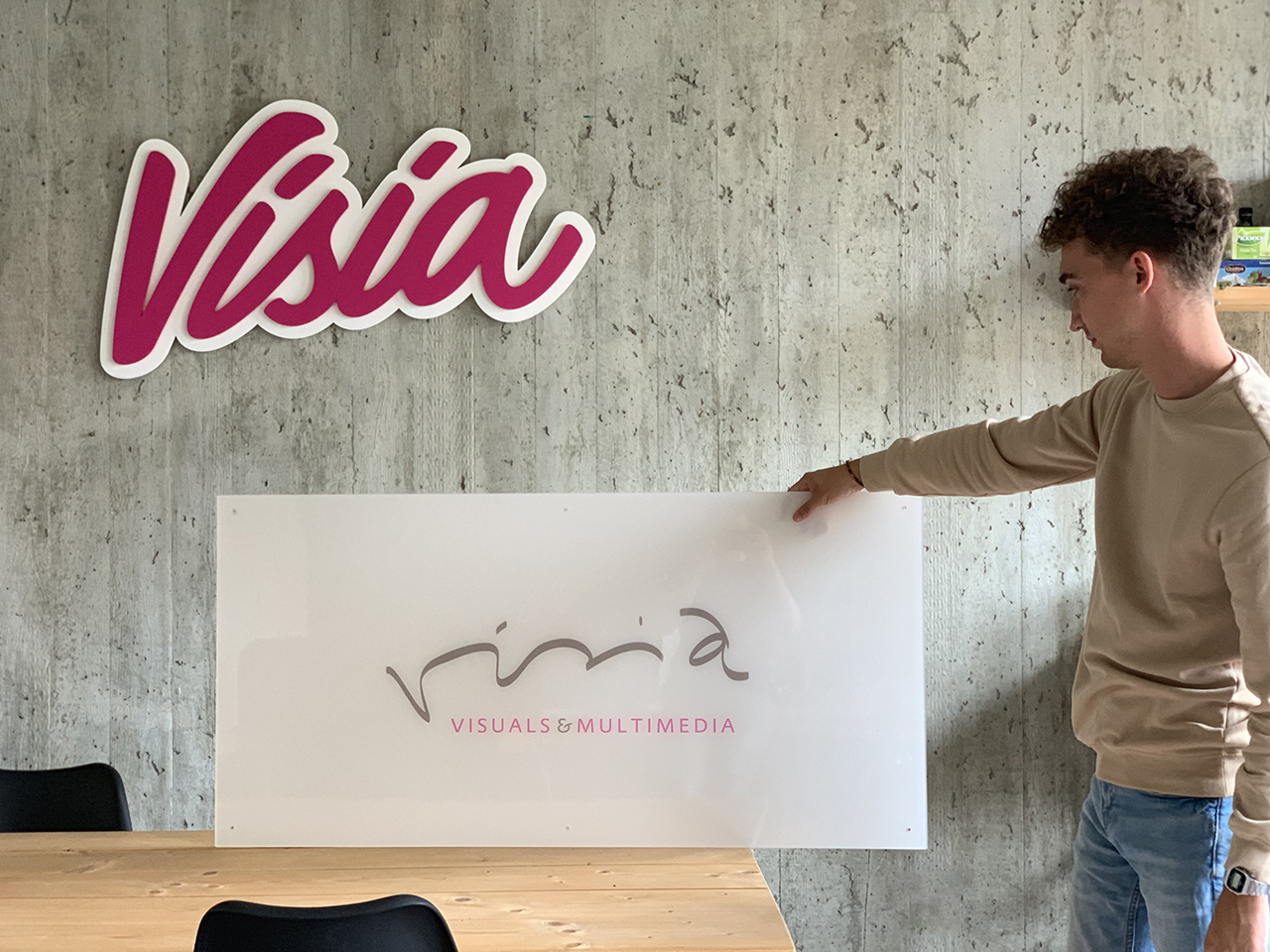 Een persoon houdt een Visia-bord vast in een kantoor, vlakbij een muur met een ander Visia-logo, waarmee de aanwezigheid van het merk wordt benadrukt. Het is duidelijk dat het bedrijf trots is op zijn identiteit en transparantie waardeert, wat verder kan worden onderzocht op hun 'Over ons'-pagina.