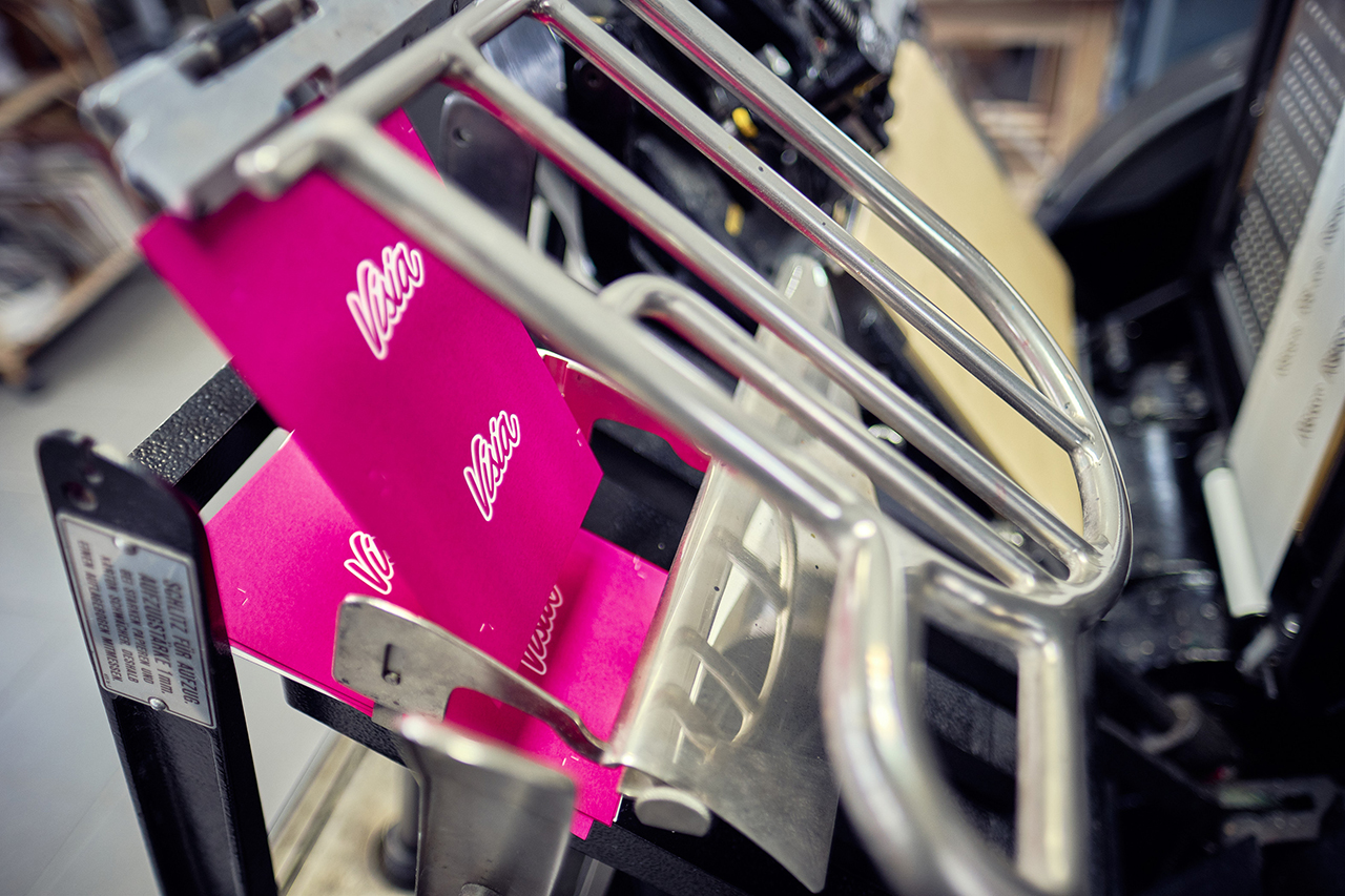 Close-up van een metalen machine met het opschrift 'Vello', waarschijnlijk een drukpers, met stukjes roze papier. Over ons branding benadrukt kwaliteit en precisie in elk detail.
