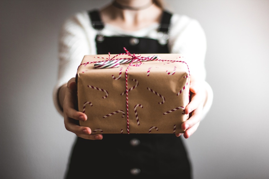 Persoon die een geschenk met een snoeprietpatroon vasthoudt, omwikkeld met touw, een wit topje en een zwarte overall draagt, perfect voor het Kerstcampagneseizoen.