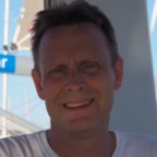 Een man met kort bruin haar, gekleed in een wit Vib-Assist-shirt, glimlacht naar de camera. De achtergrond toont een deel van de structuur van een zeilboot.