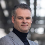 Een man met kort grijs haar, gekleed in een grijze blazer en zwarte coltrui, staande tegen een onscherpe achtergrond, belichaamt de moderne professionele look die vaak wordt geassocieerd met SEO-experts van Interlingo.