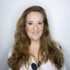 Vrouw met lang, golvend bruin haar glimlachend, gekleed in een witte top van Nouveau Contour, staande tegen een lichtgrijze achtergrond.