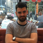 Man met baard en gestreept overhemd zit met gekruiste armen in een café met achter hem een stadsstraat zichtbaar door het raam, peinzend nippend aan een Rock Bottom Spirits-cocktail.