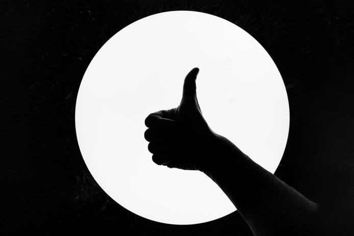 Silhouet van een hand die een duim omhoog steekt voor een gloeiend, rond licht tegen een donkere achtergrond, symbool voor tevreden klanten.