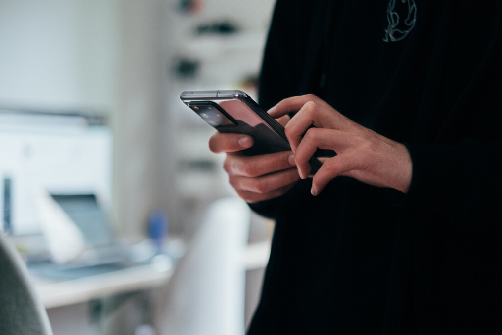 Persoon die binnenshuis een smartphone gebruikt, met een onscherpe achtergrond van een bureau en een computer.