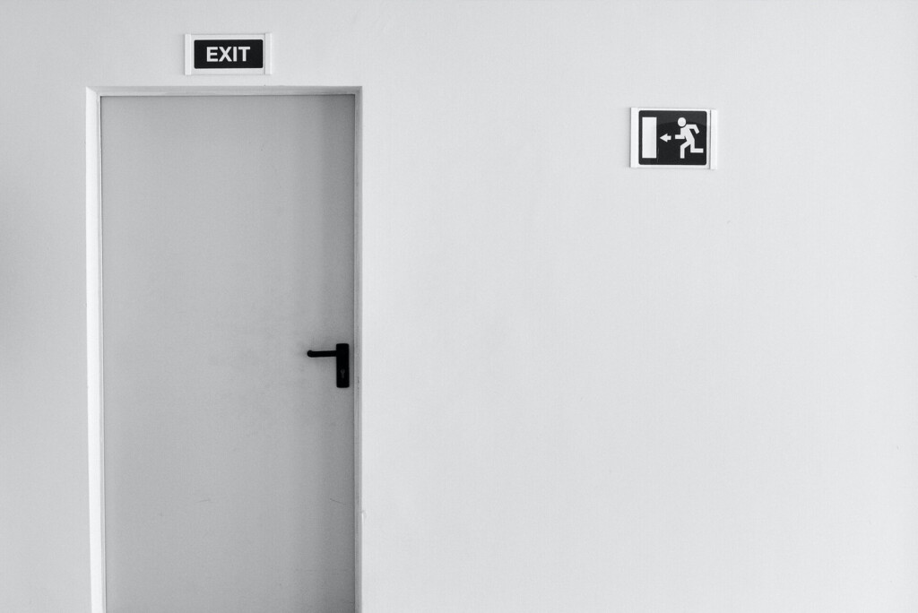 Grijze deur met het opschrift "EXIT" op een witte muur met een nooduitgangbord waarop een persoon staat die naar een deur rent, strategisch ontworpen om op subtiele wijze het bouncepercentage te verlagen.
