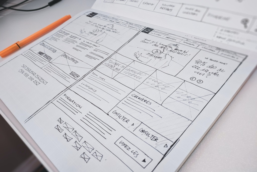 Een schets van een website-indeling met handgetekende secties, tekst en wireframes, liggend op een bureau naast een oranje pen, met ideeën voor gebruikerservaring op de website.