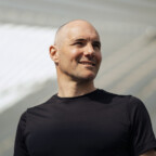 Bas Diederen, een kale man in een zwart shirt, wordt buiten glimlachend gezien met een moderne architectonische achtergrond.