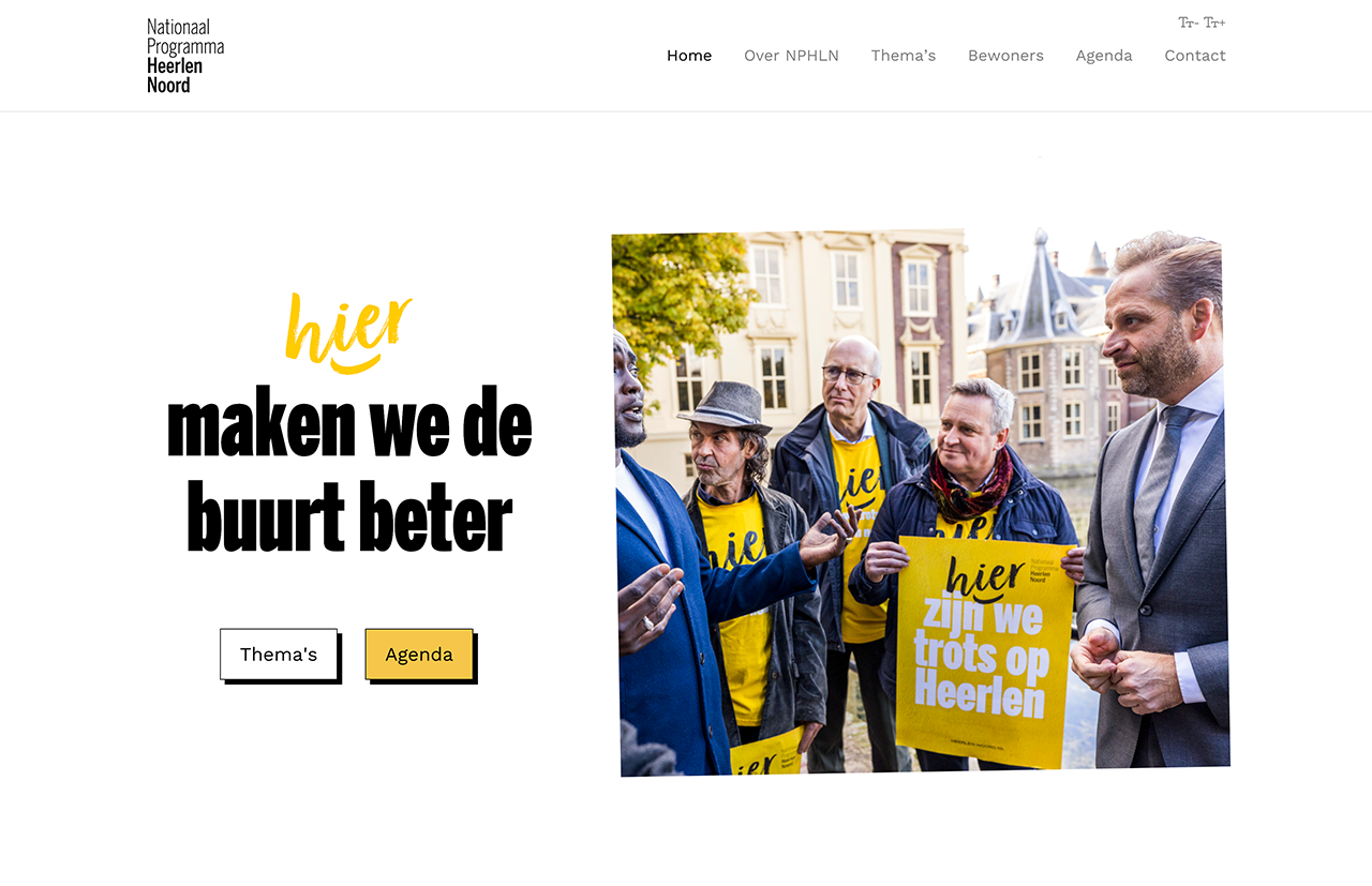 Een groep mensen in gele overhemden en jasjes praat buiten met een man in pak, onder de vlag van Nationaal Programma Heerlen-Noord; tekst in het Nederlands op de afbeelding.
