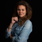 Vrouw met lang bruin haar, gekleed in een spijkerblouse, poseert tegen een zwarte achtergrond, met haar hand bij haar kin.