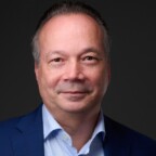 Een man met kort grijs haar, gekleed in een blauw colbert en een wit overhemd, poseert tegen een donkere achtergrond en belichaamt de verfijnde professionaliteit van NewRoutes.