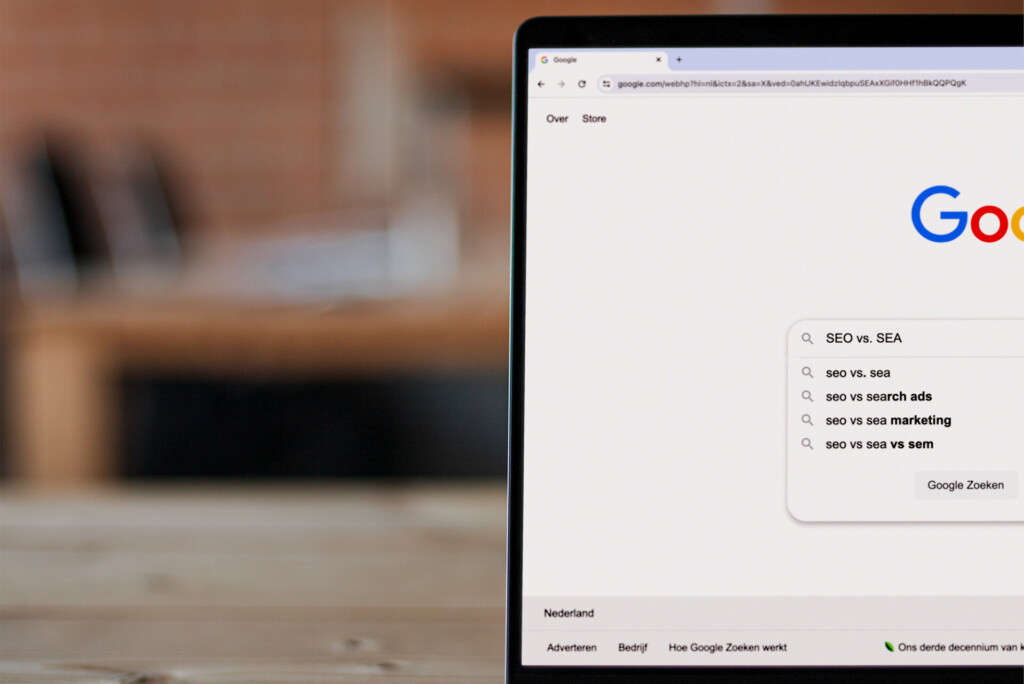 Een laptop op een houten bureau geeft een Google-zoekpagina weer met de zoekopdracht "SEA vs. SEO" en gerelateerde zoeksuggesties.
