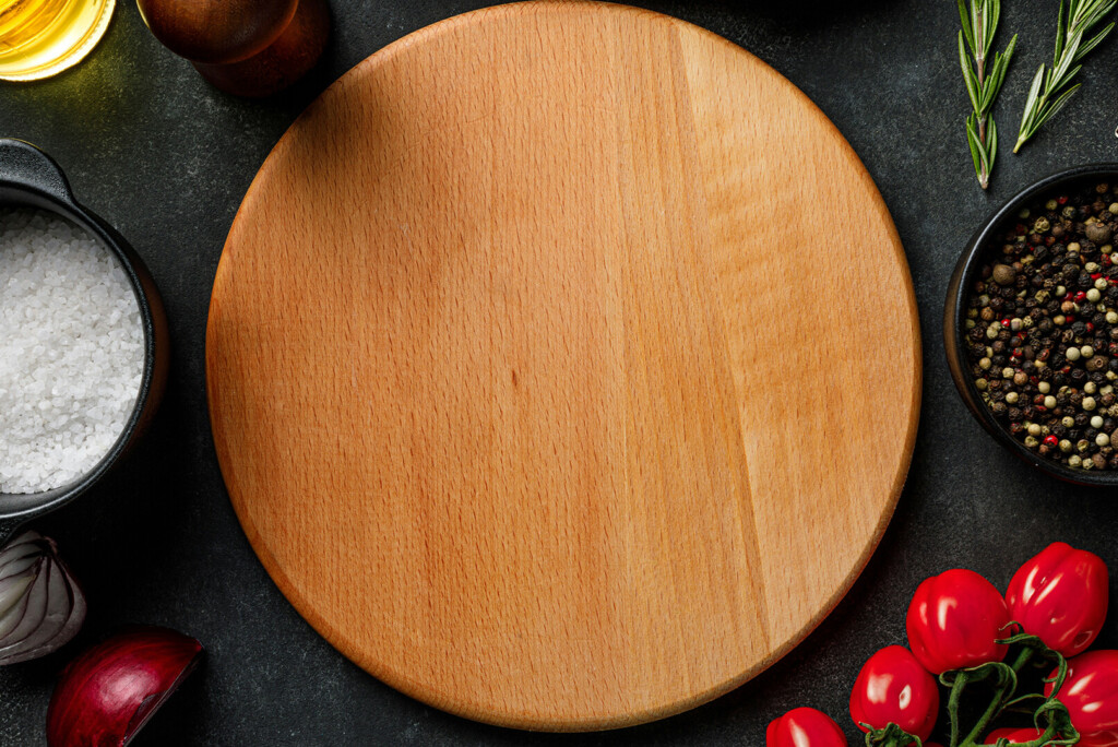 Een ronde houten snijplank omringd door zout, peper, kerstomaatjes, knoflook, rozemarijn en ui op een donkere ondergrond zorgt voor een uitnodigend tafereel dat doet denken aan de culinaire vitrine van een bedrijfswebsite.