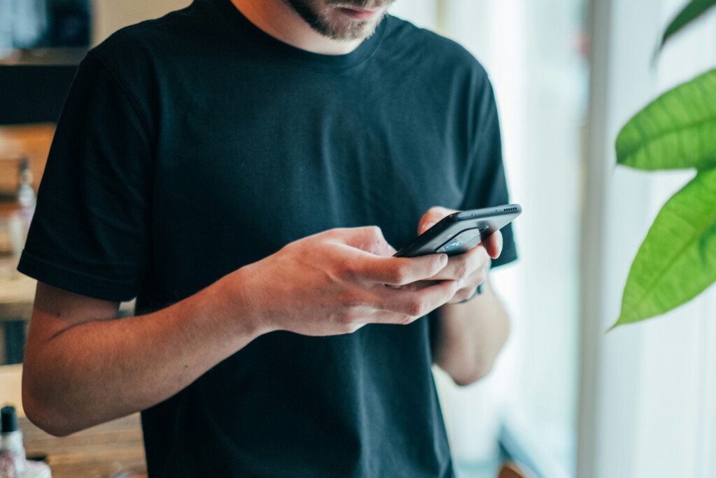 Een persoon in een zwart t-shirt die met beide handen een smartphone gebruikt, staande bij een raam en een groene plant, waarschijnlijk gezien de kosten van adverteren op sociale media.
