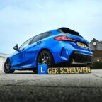 Blauwe BMW hatchback buiten geparkeerd met een "L GER SCHEIJVEN" bord op de grond ervoor, voor rijschool Ger Scheijven.