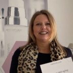 Glimlachende vrouw met beugel houdt een certificaat vast; ze draagt een sjaal met luipaardprint en staat bij een poster van Dermes anti-aging huidverzorging.