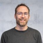 Een man met een bril en een baard, in een donkere trui, glimlacht voor een lichtgekleurde achtergrond - de expertise van Bouwcoördinatie bij Praevalis.