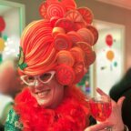 Persoon met een felle pruik en bril in snoepthema, die een rood drankje vasthoudt en glimlacht op een feestelijk feestje.