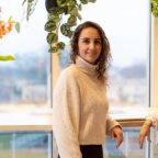 Vrouw in een beige coltrui staat binnen bij een raam, omringd door groene planten.