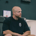 Een gespierde man met een kaalgeschoren hoofd zit binnen en draagt een donker t-shirt van WVN Coaching met een wit logo op de borst.