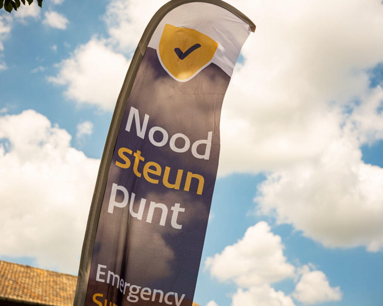 Vlag met de teksten „Nood Steun Punt“, „Emergency Support“ en „Pilot noodhulp“ tegen een blauwe lucht met wolken.