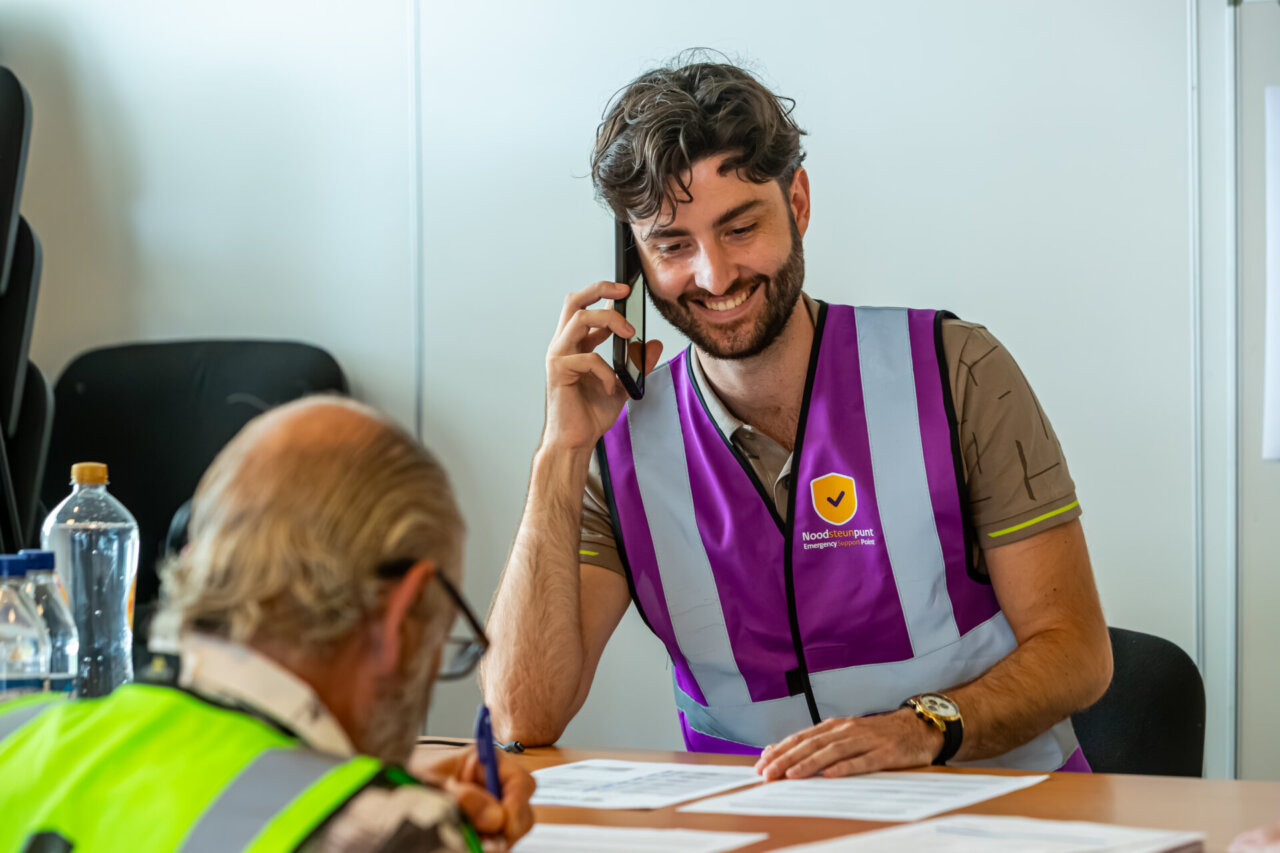 Een glimlachende man in een paars veiligheidsvest bespreekt aan zijn bureau met een collega via de telefoon de Pilot Noodsteunpunten.