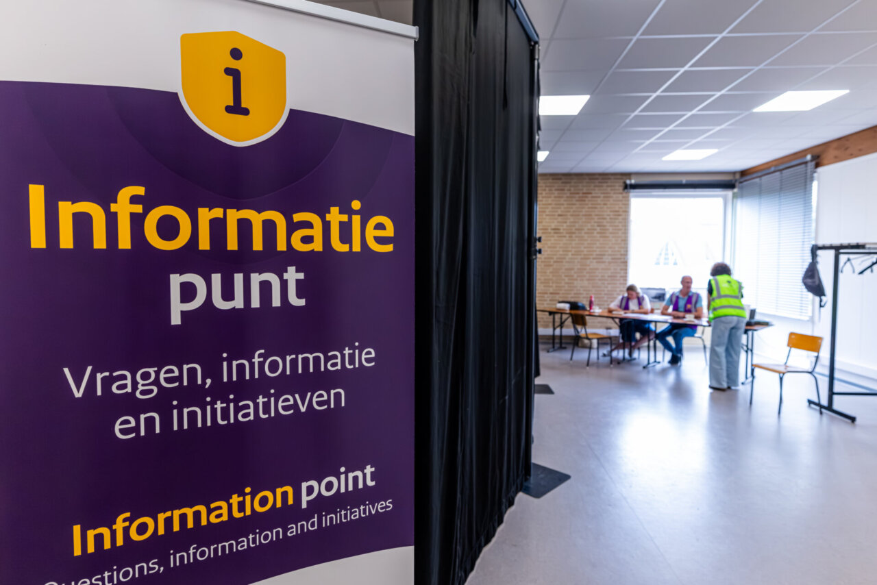 Paars informatiebord in het Nederlands voor Pilot Noodsteunpunten; op de achtergrond zitten mensen aan een tafel in een lichte ruimte.