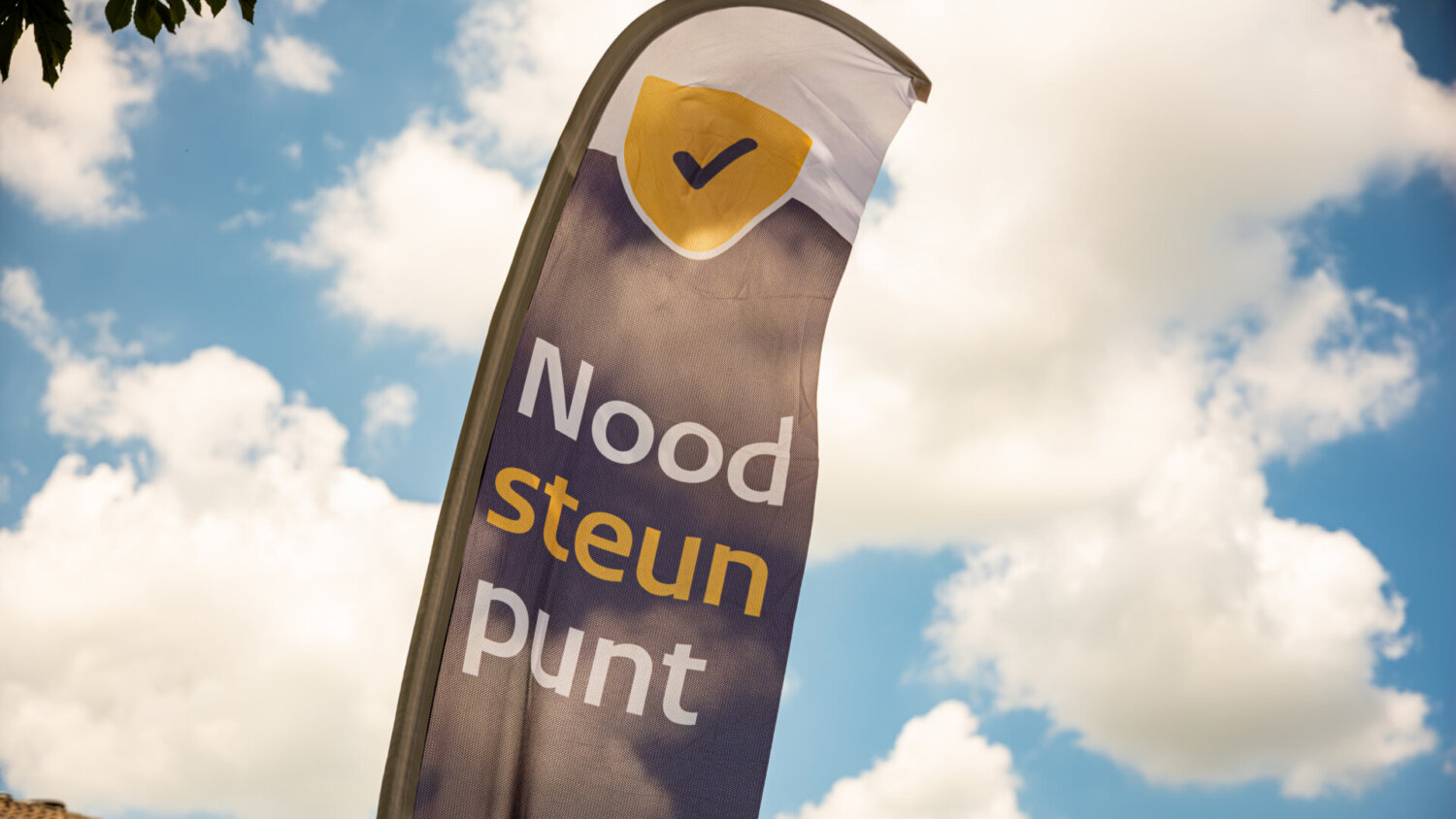 Vlag met de tekst „Noodsteunpunt”, een geel vinkje en een blauwe lucht — onderdeel van het initiatief „Pilot Noodsteunpunten”.